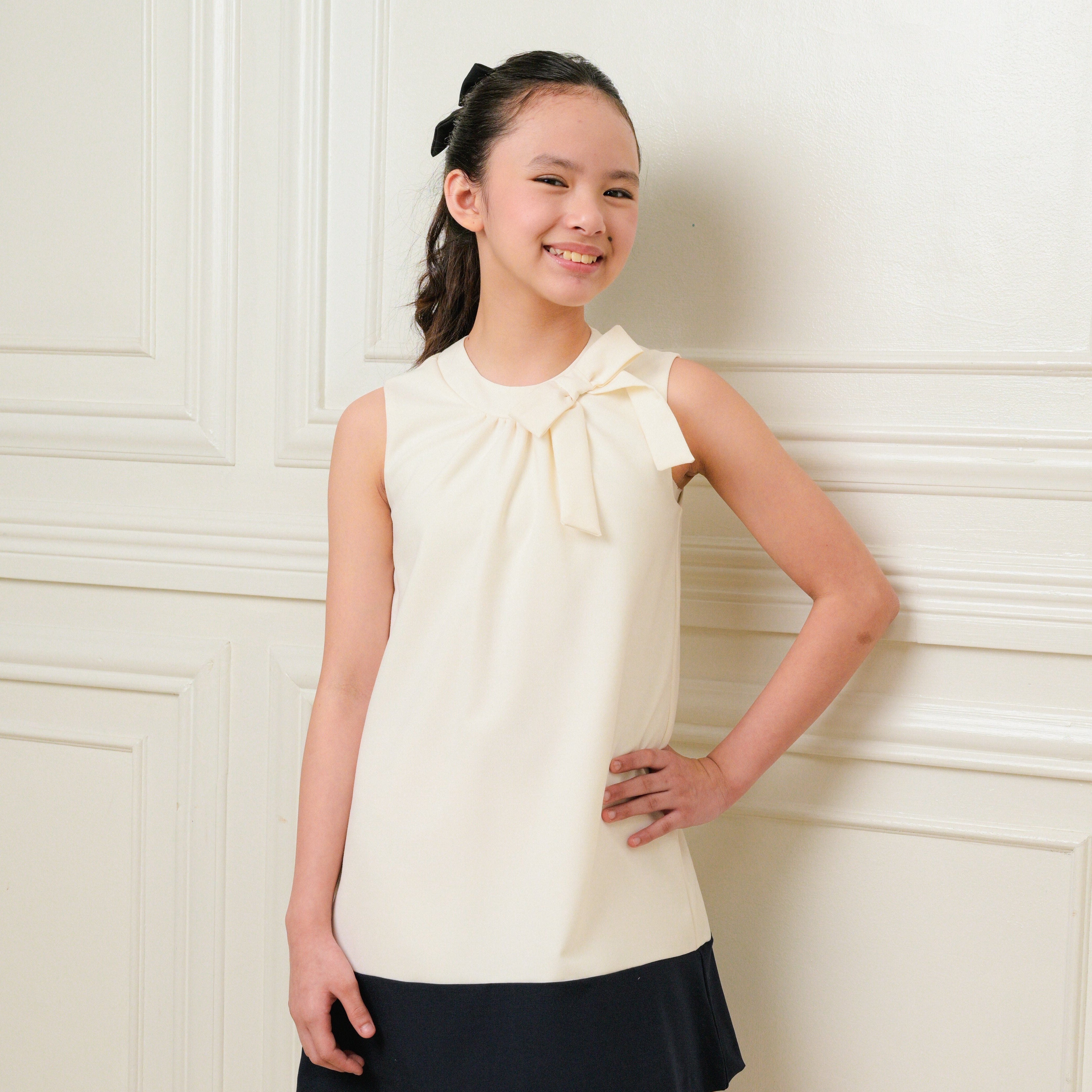 Young girl wearing a white blouse with a bow and black skirt against a white wall.
