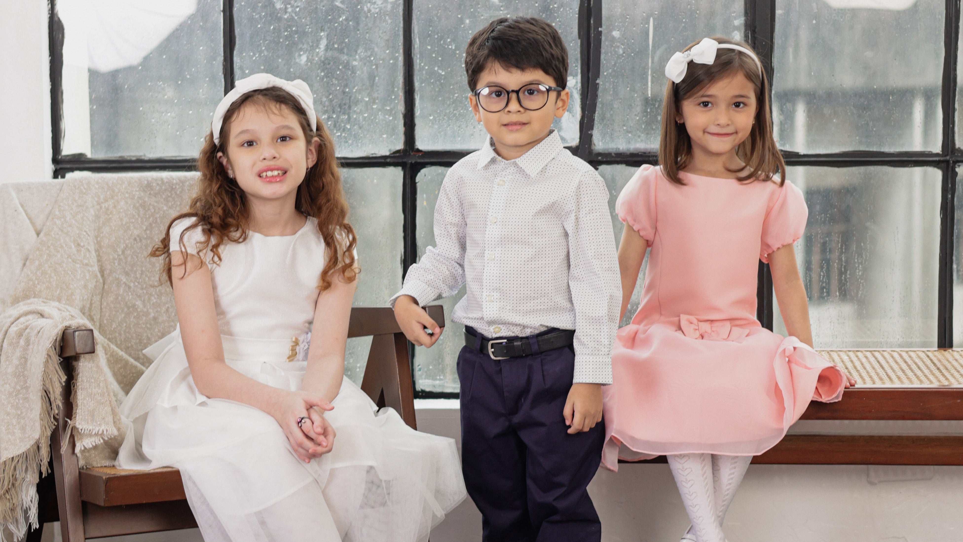 Three children wearing formal party dresses and suits pose together in front of a large black-framed window. The girl on the left wears a white sleeveless dress with a tulle skirt, the boy in the center wears a white shirt and dark trousers, and the girl on the right wears a pink dress.